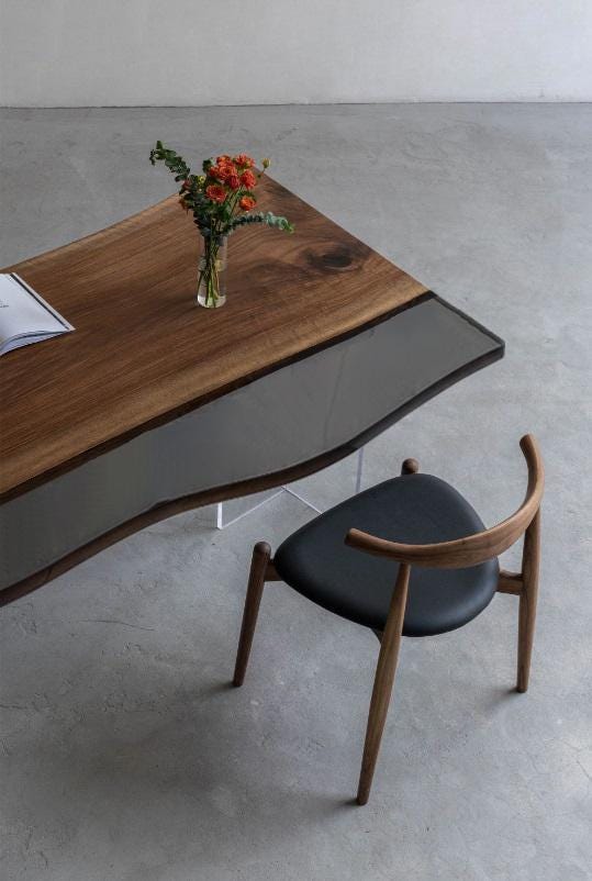 A wooden desk with a live edge and a gray glass insert, featuring a clear glass vase with orange flowers and a book on top. Beside the desk is a wooden chair with a black upholstered seat and a curved backrest.