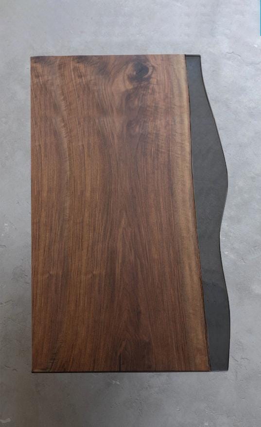 A rectangular table top featuring a natural wood grain surface with a rich brown finish on the left side and a contrasting dark gray, smooth, wavy-edged material panel attached along the entire right side of the piece.