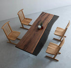 A rectangular dining table featuring a natural live-edge wood slab paired with a smooth black section, supported by clear acrylic legs. Four light-toned wooden chairs with spindle backs and angled bases are arranged around the table.