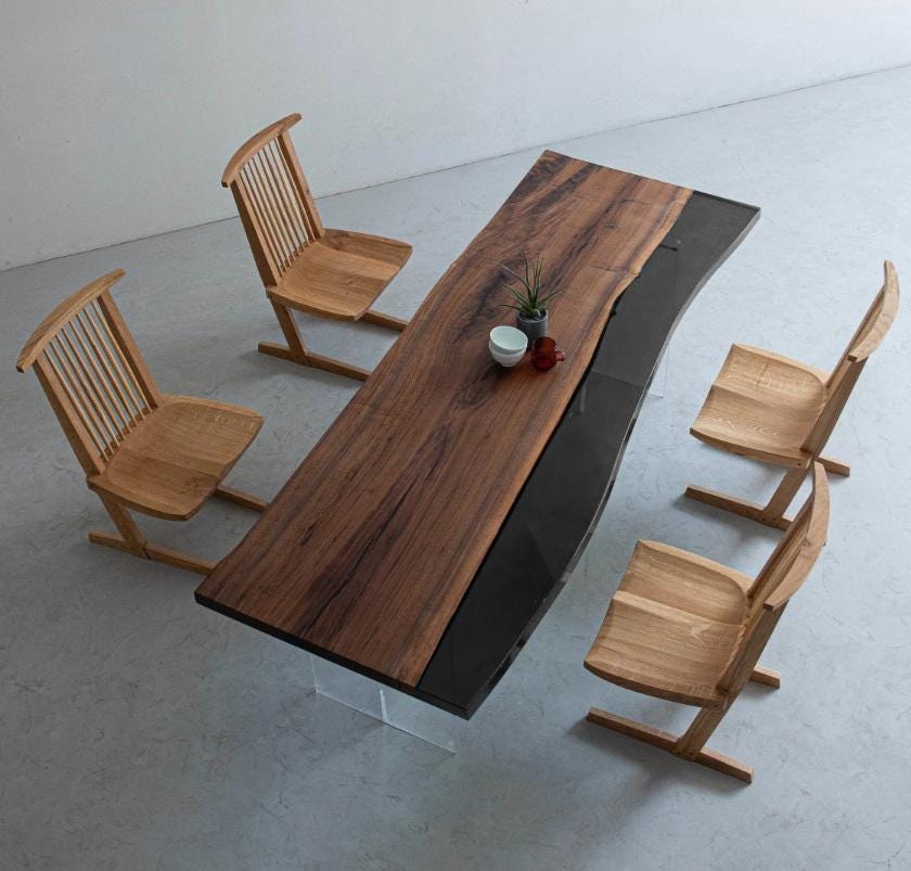 A rectangular dining table featuring a natural live-edge wood slab paired with a smooth black section, supported by clear acrylic legs. Four light-toned wooden chairs with spindle backs and angled bases are arranged around the table.