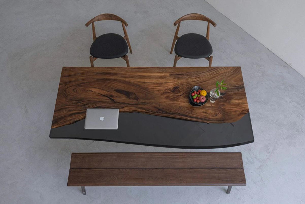 A rectangular dining table featuring a natural live-edge wood slab on one side and a smooth, solid black resin section on the other. The set includes two wooden chairs with black upholstered seats and a matching wooden bench.