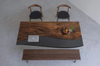 A rectangular dining table featuring a natural live-edge wood slab on one side and a smooth, solid black resin section on the other. The set includes two wooden chairs with black upholstered seats and a matching wooden bench.
