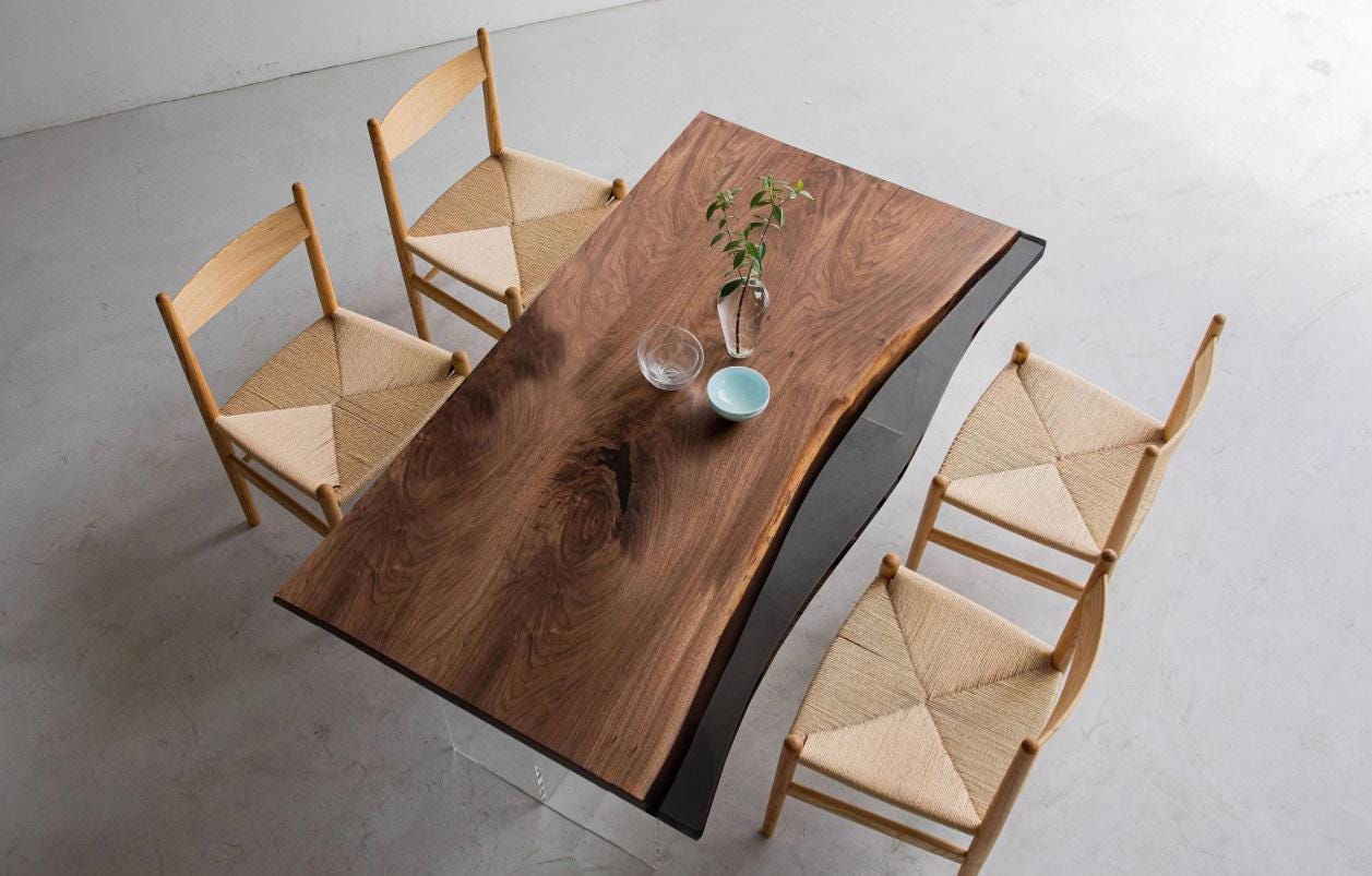 A rectangular dining table with a natural live-edge dark wood top and a black side panel, supported by clear acrylic legs. Four light-colored wooden chairs with woven seats are arranged around the table, which holds a glass vase.