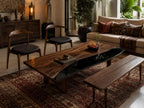 A rectangular coffee table featuring a natural live-edge wood slab surface with a dark, glossy black resin inlay running through the center, supported by sturdy wooden legs, positioned in a room with two chairs and a wooden bench.