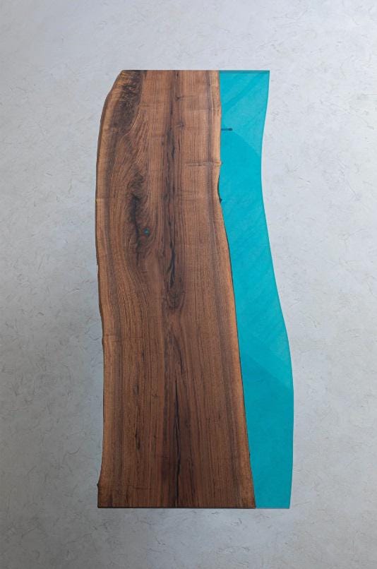 A rectangular table top featuring a natural live edge brown wood slab on one side and a smooth, translucent turquoise blue resin section on the other, creating a contrasting wavy border between the organic wood grain and the resin.