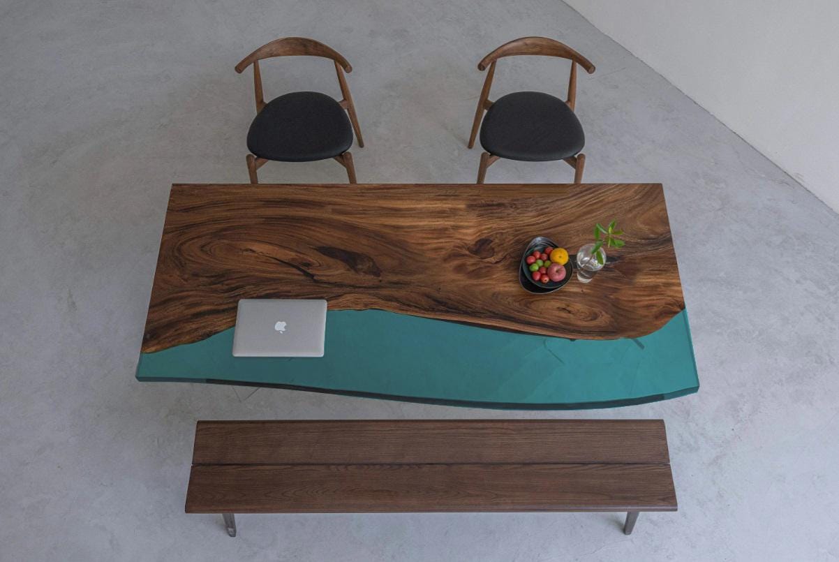 A rectangular dining table featuring a natural live-edge dark wood slab paired with a smooth, translucent teal resin section. The set includes two wooden chairs with black upholstered seats and a matching long wooden bench.