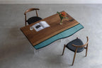 A rectangular dining table featuring a natural wood grain surface with a wavy teal resin inlay, supported by clear acrylic legs. Two wooden chairs with black upholstered seats are positioned on opposite sides of the table.