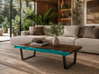 A rectangular coffee table featuring a natural dark wood slab with a live edge and a translucent turquoise resin inlay. The table is supported by two black metal U-shaped legs and holds a small ceramic vase and a stack of books.