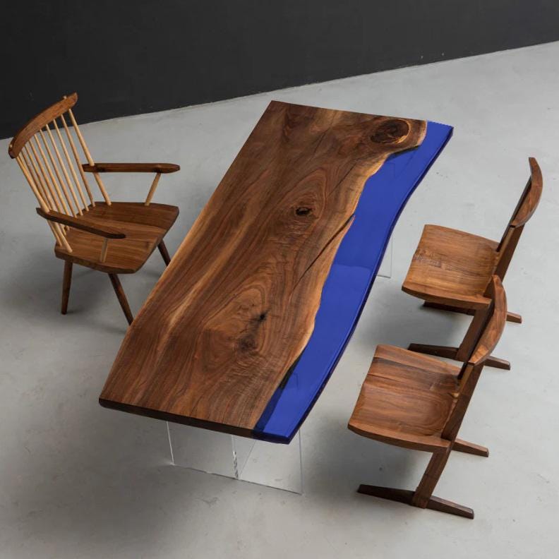 A rectangular dining table with a natural live-edge dark wood slab and a vibrant blue resin inlay, supported by clear acrylic legs. The set includes one spindle-back wooden chair and two matching wooden chairs with angled legs.