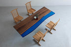A rectangular dining table featuring a natural wood grain surface with a live edge and a vibrant blue resin insert, supported by clear acrylic legs, accompanied by four light-colored wooden chairs with spindle backs.