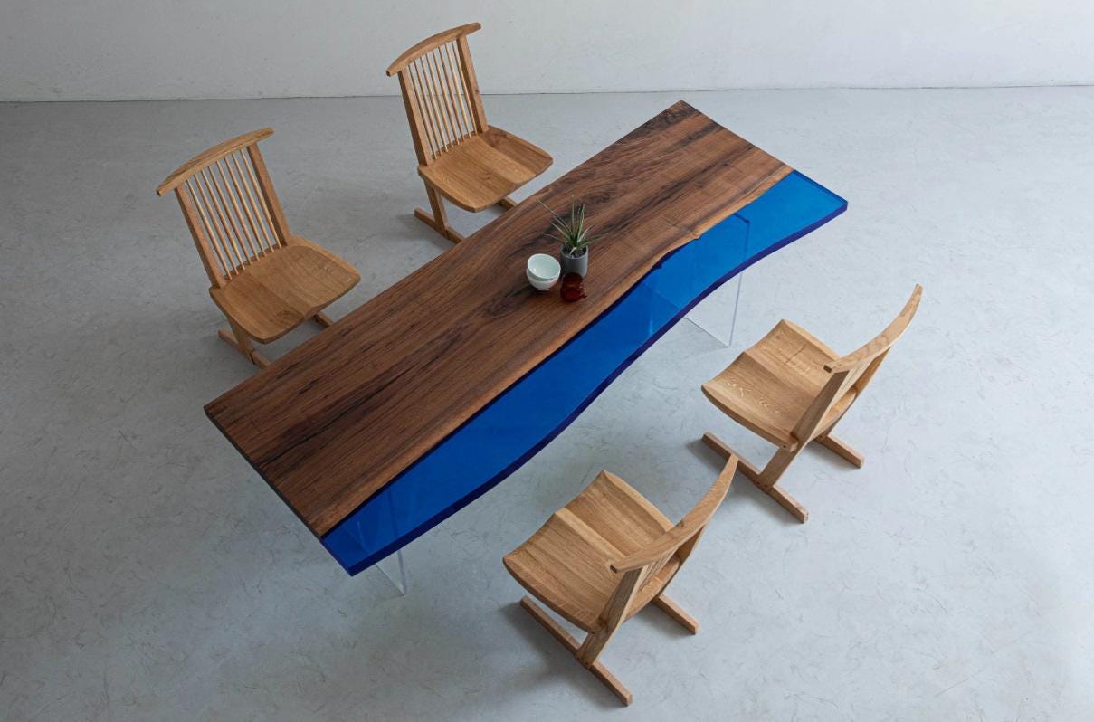 A rectangular dining table featuring a natural wood grain surface with a live edge and a vibrant blue resin insert, supported by clear acrylic legs, accompanied by four light-colored wooden chairs with spindle backs.
