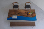 A rectangular dining table featuring a natural live-edge wood slab with a prominent dark brown grain and a smooth, solid blue resin section. The table is accompanied by two wooden chairs with black seats and a matching wooden bench.
