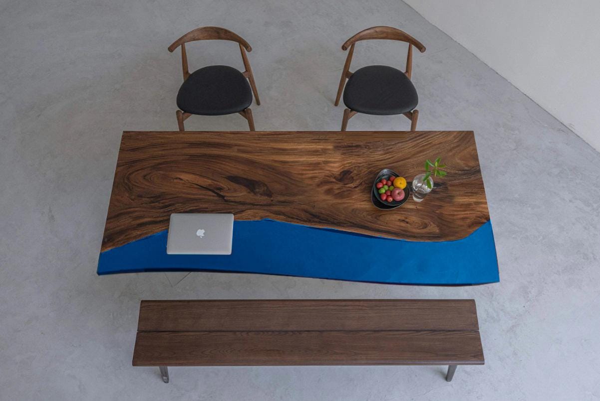 A rectangular dining table featuring a natural live-edge wood slab with a prominent dark brown grain and a smooth, solid blue resin section. The table is accompanied by two wooden chairs with black seats and a matching wooden bench.