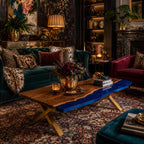 A rectangular coffee table featuring a natural wood slab with a live edge and a vibrant blue resin inlay. The table is supported by a gold-colored metal X-frame base. The surface holds a gold bowl filled with dark flowers.