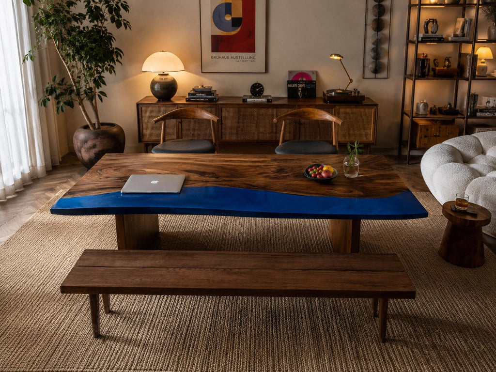 A rectangular dining table featuring a natural wood grain surface with a prominent, smooth, vibrant blue resin inlay running along one side. The table is supported by two solid wood block bases and is accompanied by a matching wooden bench.