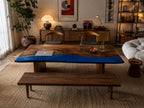 A rectangular dining table featuring a natural wood grain surface with a prominent, smooth, vibrant blue resin inlay running along one side. The table is supported by two solid wood block bases and is accompanied by a matching wooden bench.
