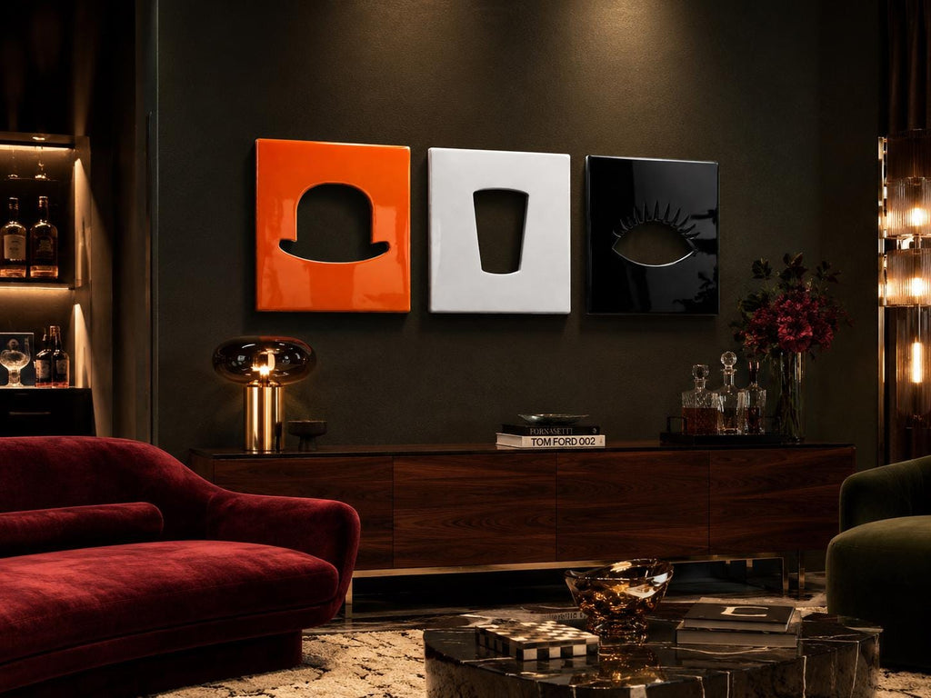 Three square wall art panels featuring cutouts of a bowler hat, a drinking glass, and an eye. The panels are colored orange, white, and black respectively, with a glossy finish, mounted horizontally on a dark wall above a wooden cabinet.