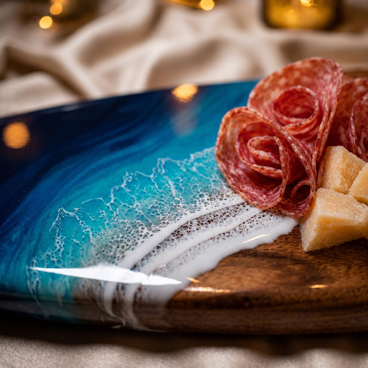 A wooden serving board featuring a glossy resin inlay that mimics ocean waves with deep blue and turquoise swirls, accented by white foam details. The board is topped with folded slices of cured salami and cubes of hard cheese.