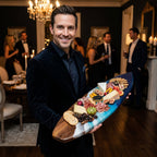 A surfboard-shaped serving board made of dark wood with a blue and white resin ocean-inspired design, holding an assortment of crackers, various cheeses, sliced meats, fresh figs, kiwi slices, and a small bowl of black caviar.