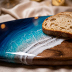 A wooden serving board featuring a decorative resin inlay that mimics ocean waves. The design transitions from deep blue to turquoise, with white lacy foam details separating the resin from the natural brown wood grain surface.