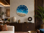 A round wall clock featuring a coastal ocean scene with blue water, white sea foam, and a sandy beach. The clock has a silver frame, silver Roman numerals, and silver hands centered on the face, depicting a serene beach landscape.