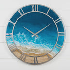 A round wall clock featuring a silver metal frame with Roman numerals. The clock face displays a realistic ocean scene with deep blue water, white crashing waves, and a sandy beach shoreline. Two silver hands indicate the time.