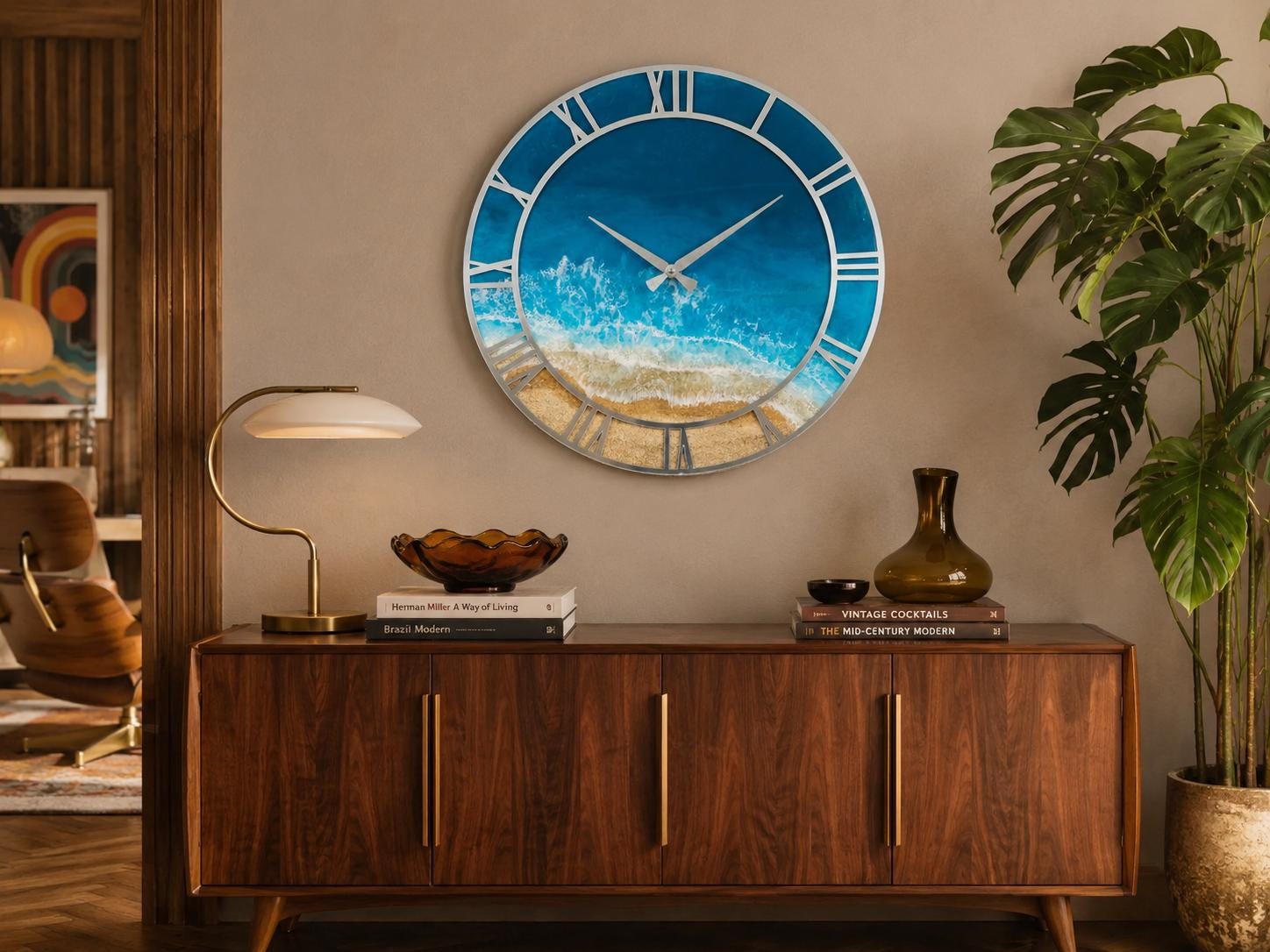 A large circular wall clock featuring a blue ocean wave design with white sea foam and a sandy beach shoreline. The clock has a silver frame, silver hands, and large silver Roman numerals positioned around the outer edge.