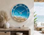 A round wall clock featuring a silver frame and Roman numerals. The clock face displays a detailed resin art depiction of a sandy beach meeting turquoise ocean waves with white sea foam, complemented by two slender silver clock hands.