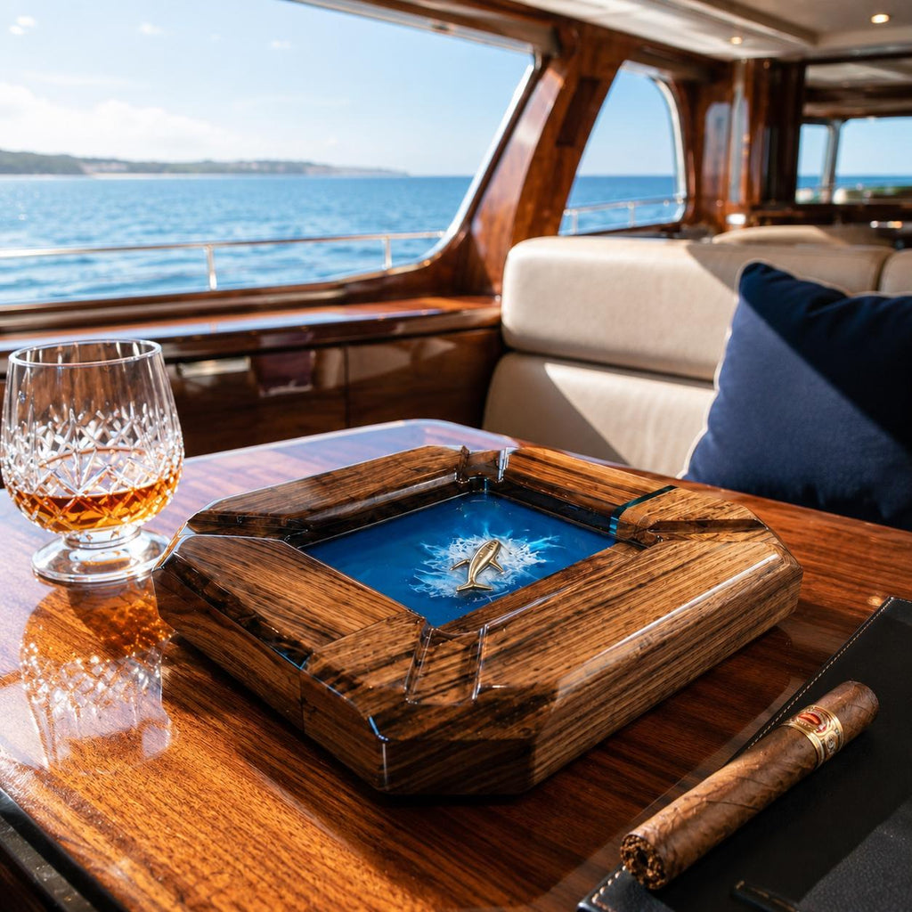 A square wooden ashtray with a polished dark grain finish features a central blue resin inlay depicting a small metallic whale breaching the surface. A crystal glass filled with amber liquid and a brown cigar rest nearby.