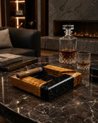 A square ashtray constructed from a combination of polished light brown wood with a prominent grain and a solid black material featuring white speckles. A single brown cigar with a gold band rests across the ashtray rim.