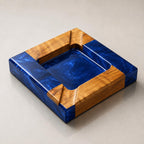A square tray featuring a combination of natural light brown wood and deep blue swirled resin. The design includes four wooden corner sections and a central recessed blue resin basin with smooth, angled edges and a polished finish.