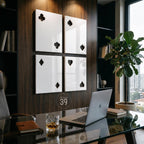 Four rectangular white wall panels featuring cut-out playing card suit symbols including clubs, spades, diamonds, and hearts. The panels are mounted on a dark wood-paneled wall above a glass desk with a laptop and a glass of amber liquid.