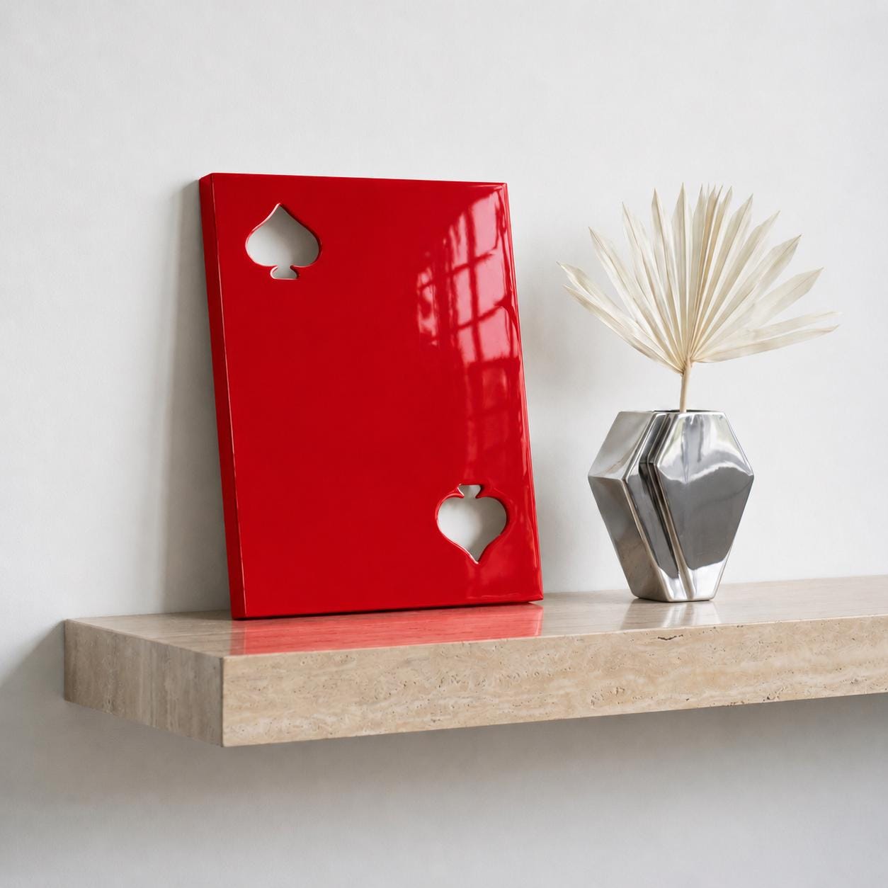A rectangular red wall art piece featuring two cutout spade symbols, one in the upper left and one in the lower right corner. Next to it sits a silver geometric vase containing a single dried, fan-shaped cream-colored palm leaf.