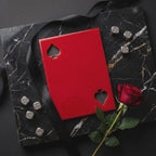 A glossy red rectangular card with two spade cutouts, accompanied by five silver dice and a single red rose. The items are arranged on a black marble surface with a black ribbon draped across the scene.