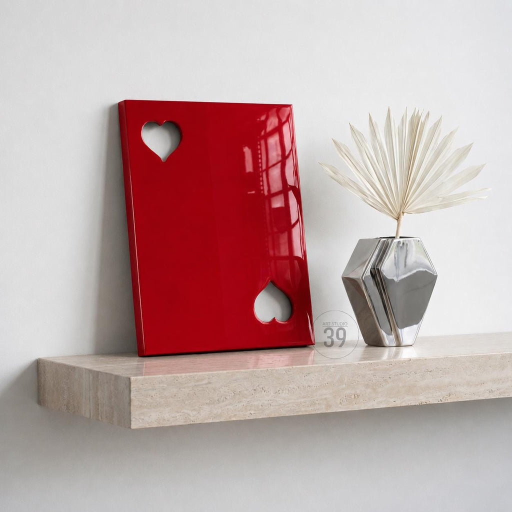 A glossy red rectangular panel featuring two heart-shaped cutouts, positioned next to a silver geometric vase containing a dried white palm leaf. The panel has a smooth, reflective surface and is displayed on a light beige stone shelf.