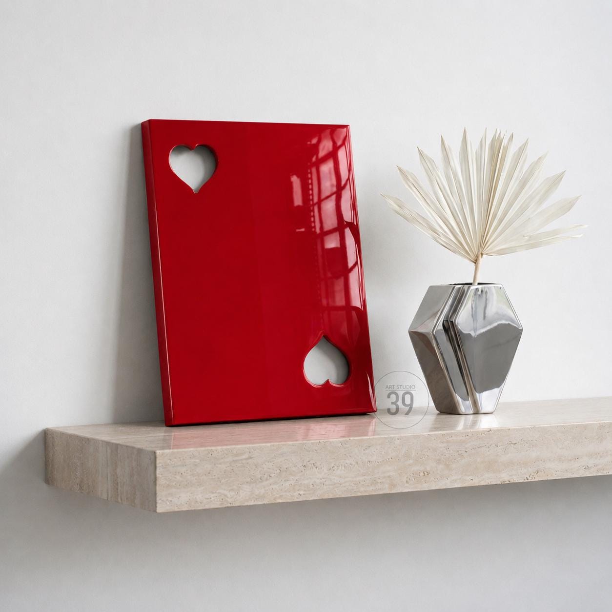 A glossy red rectangular panel featuring two heart-shaped cutouts, positioned next to a silver geometric vase containing a dried white palm leaf. The panel has a smooth, reflective surface and is displayed on a light beige stone shelf.
