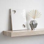 A white rectangular plaque featuring two heart-shaped cutouts, positioned next to a silver geometric vase containing a dried palm leaf. The items rest on a beige stone shelf with a visible natural texture and horizontal grain.