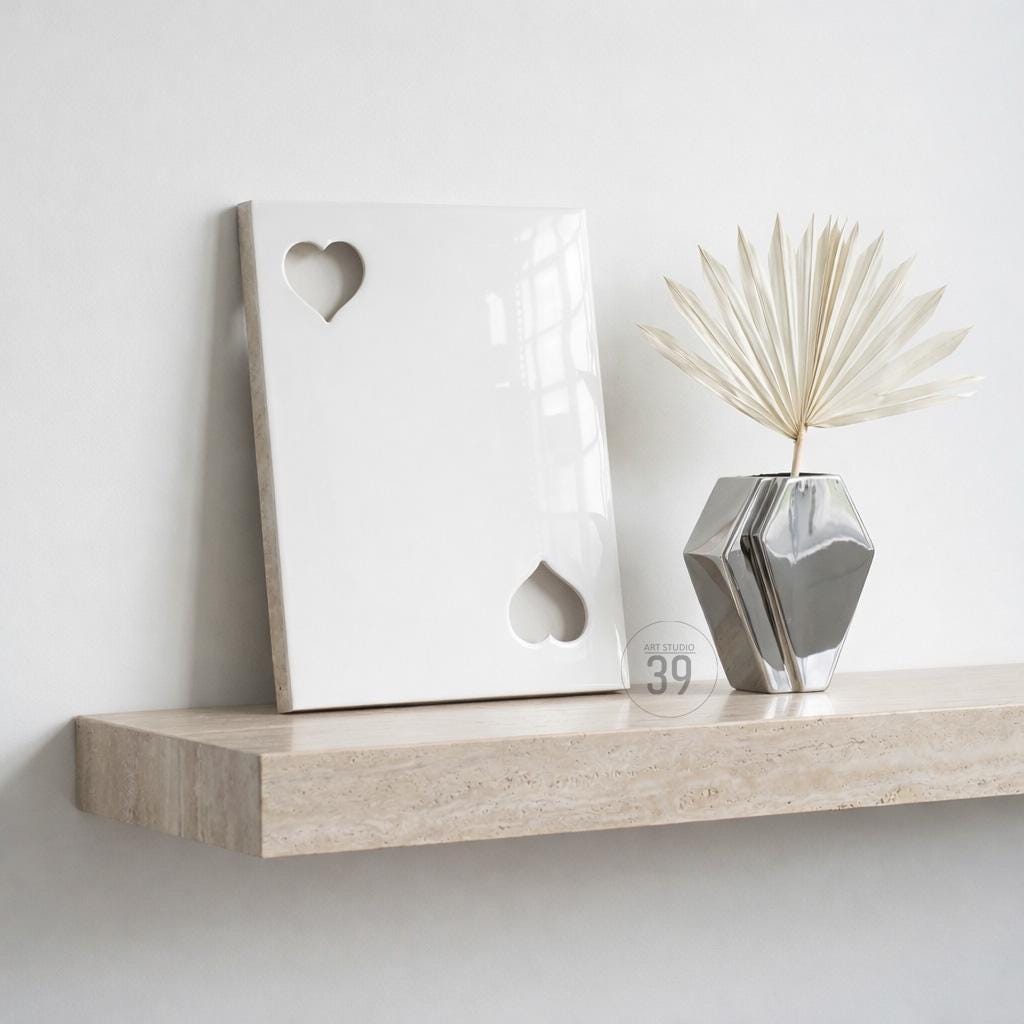 A white rectangular plaque featuring two heart-shaped cutouts, positioned next to a silver geometric vase containing a dried palm leaf. The items rest on a beige stone shelf with a visible natural texture and horizontal grain.