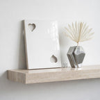 A white rectangular plaque featuring two heart-shaped cutouts, positioned next to a silver geometric vase containing a dried palm leaf. The items rest on a beige stone shelf with a visible natural texture and horizontal grain.
