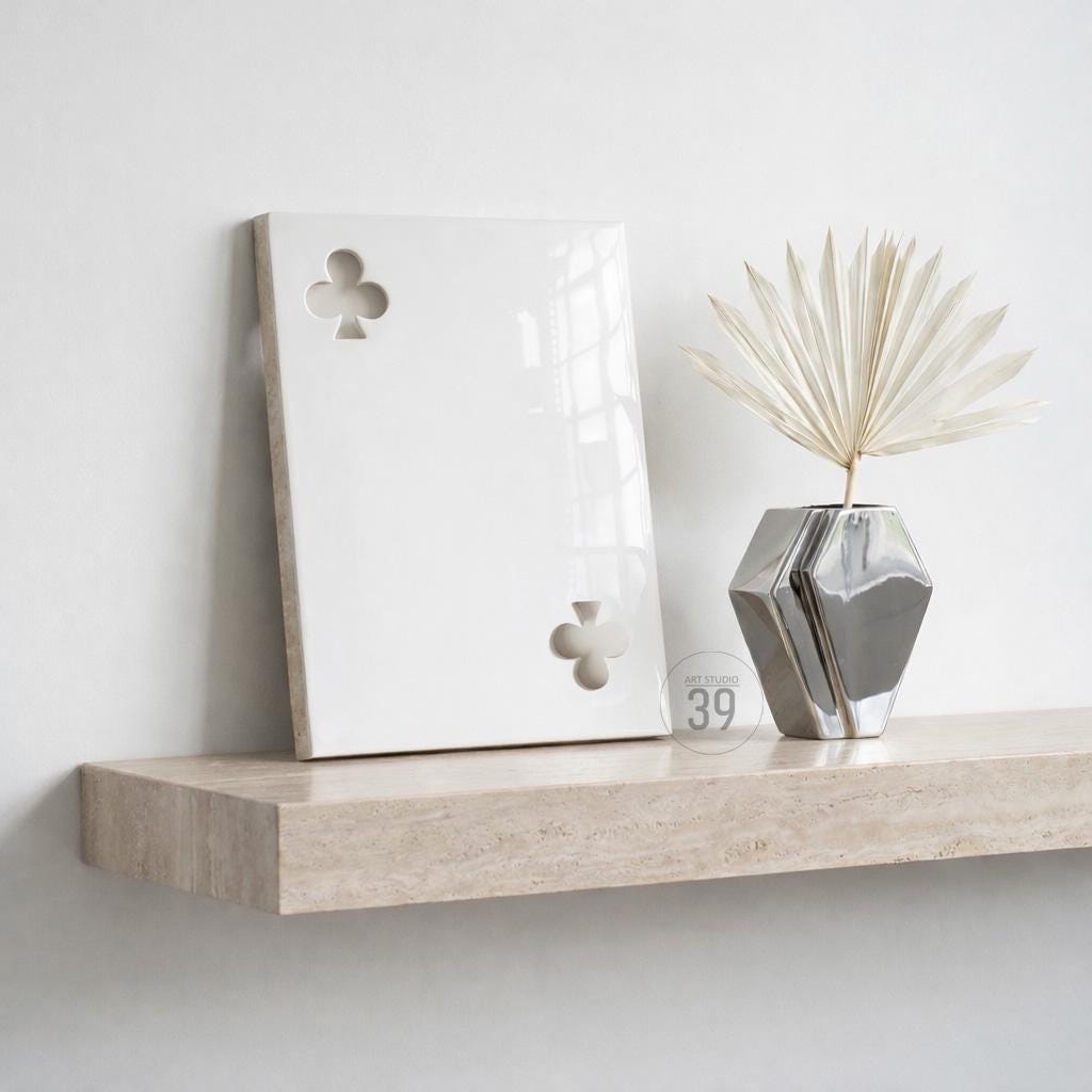 A rectangular white ceramic plaque featuring a cutout club suit symbol in the top left and bottom right corners, displayed next to a silver geometric vase containing a dried white palm leaf fan on a beige stone shelf.