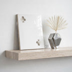 A rectangular white ceramic plaque featuring a cutout club suit symbol in the top left and bottom right corners, displayed next to a silver geometric vase containing a dried white palm leaf fan on a beige stone shelf.