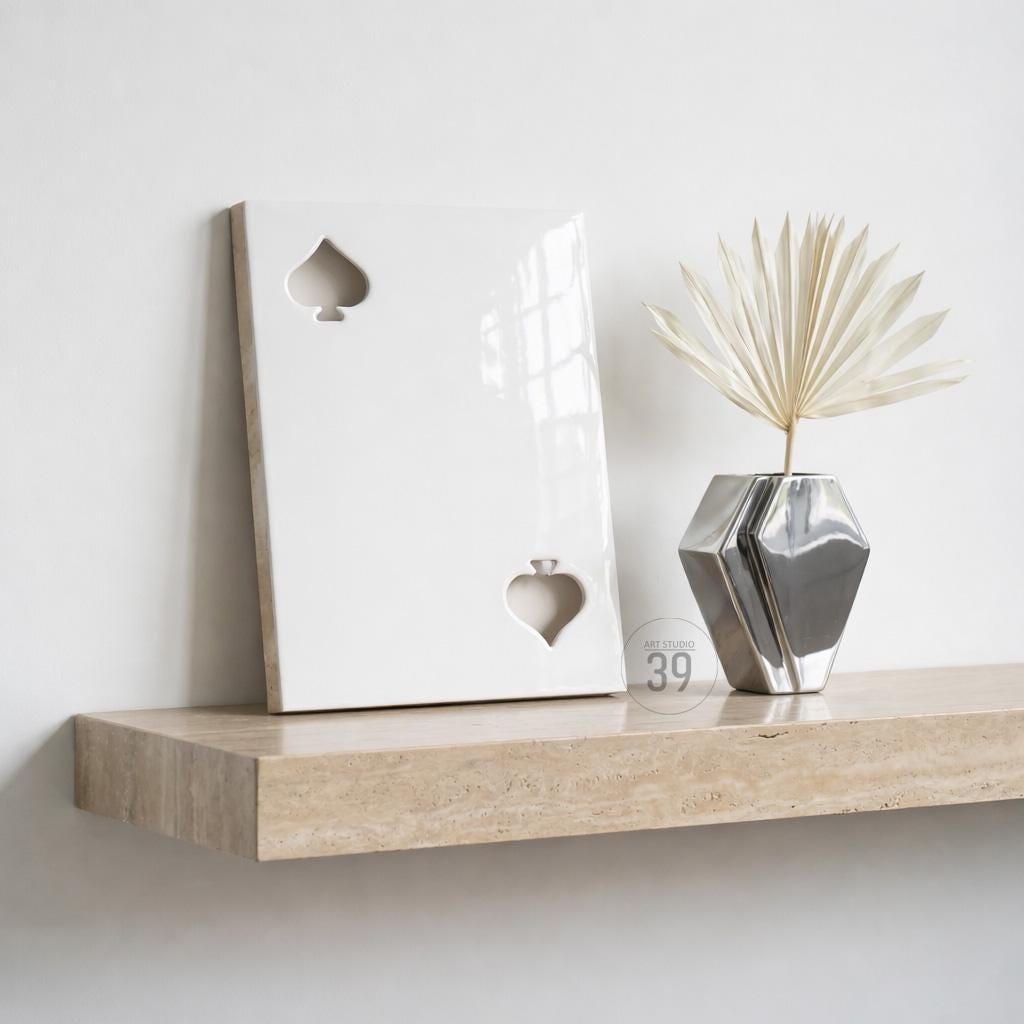 A rectangular white ceramic plaque featuring two cutout spade symbols, one in the upper left and one in the lower right corner. Beside it sits a faceted, metallic silver vase containing a single, dried, fan-shaped palm leaf.