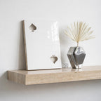 A rectangular white ceramic plaque featuring two cutout spade symbols, one in the upper left and one in the lower right corner. Beside it sits a faceted, metallic silver vase containing a single, dried, fan-shaped palm leaf.