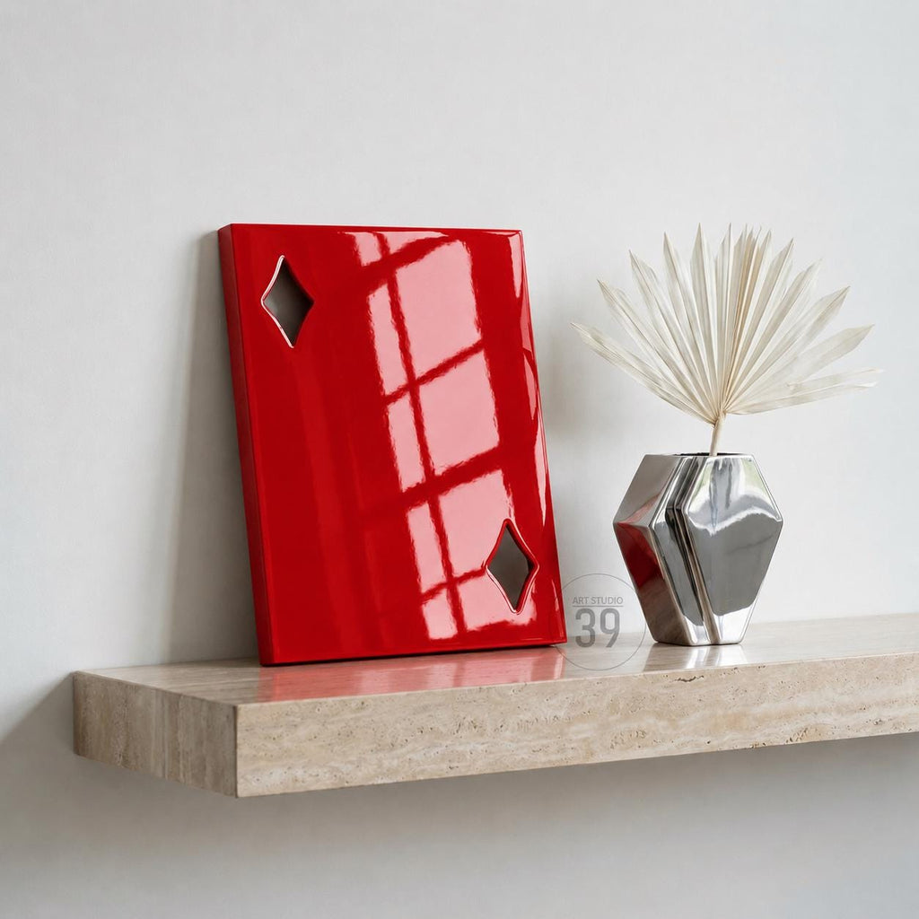 A rectangular red wall art piece featuring two diamond-shaped cutouts, positioned next to a silver geometric vase containing a white dried palm leaf fan. The red object has a glossy finish reflecting light from a nearby window.