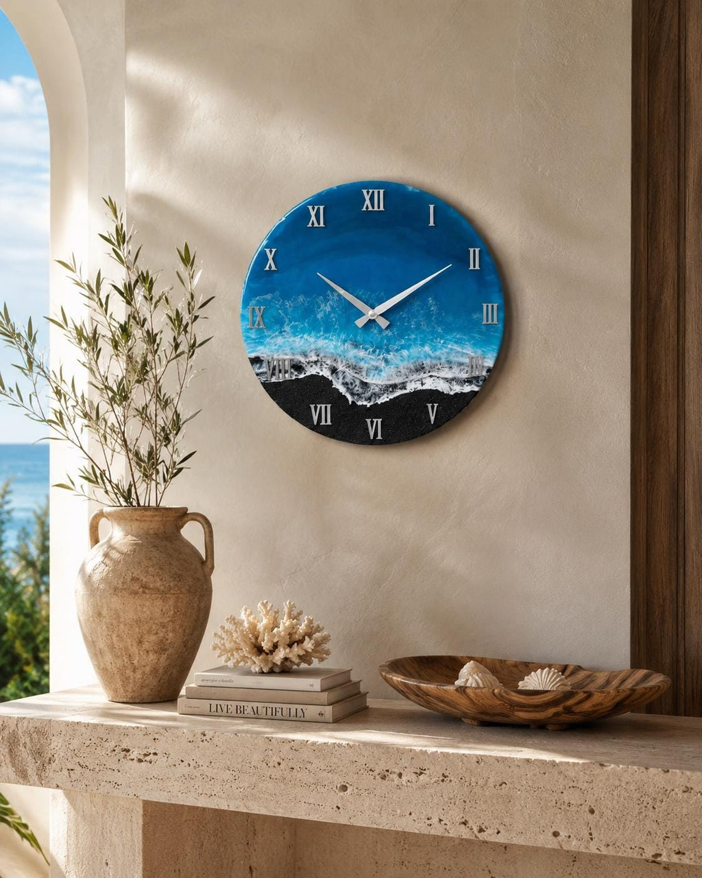 A circular wall clock featuring a resin art design that mimics a beach scene with deep blue water, turquoise waves, white sea foam, and a black sand shoreline. The clock face displays silver Roman numerals and thin silver clock hands.