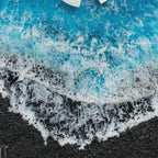 A resin art piece featuring a textured black granular base representing sand, topped with layers of vibrant blue resin and white lacy foam effects that mimic the appearance of ocean waves crashing onto a dark shoreline.