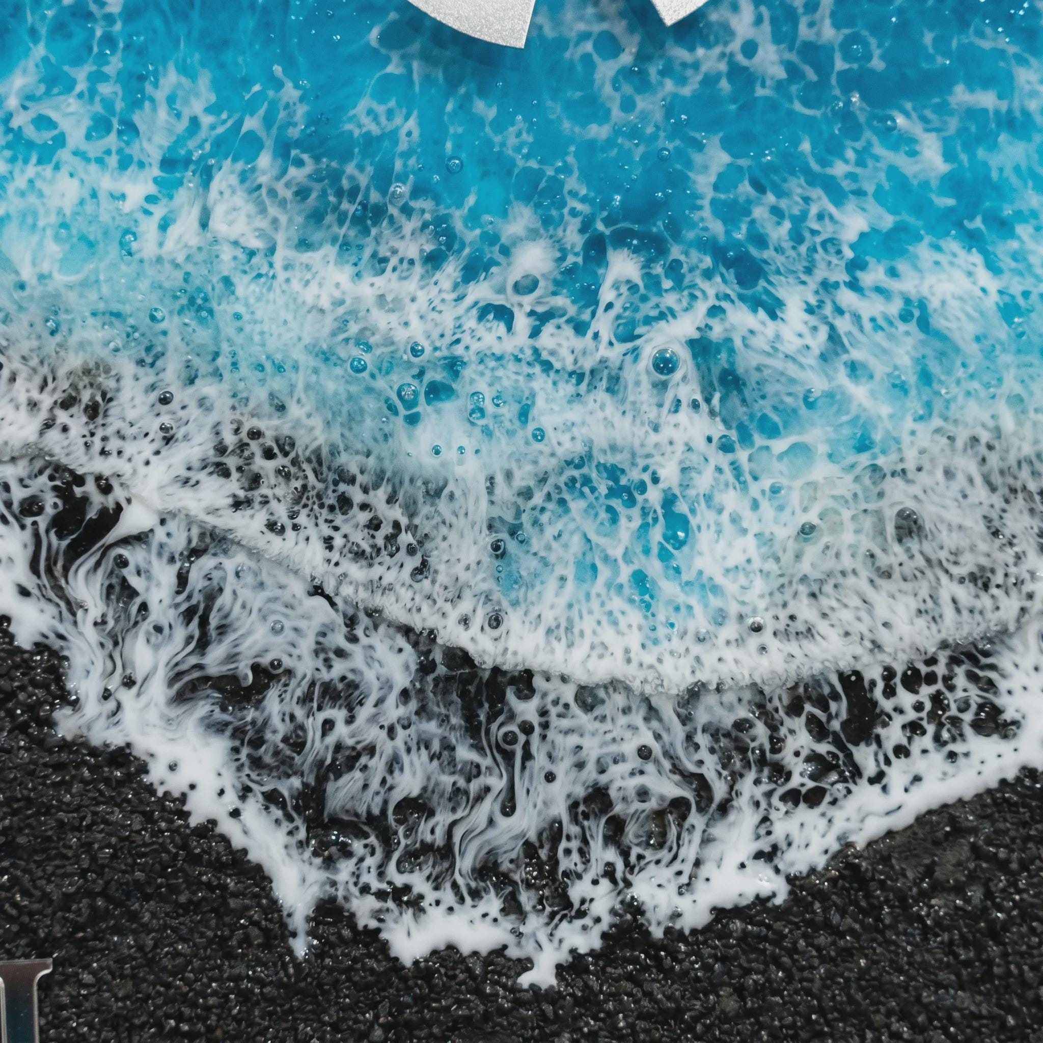 A resin art piece featuring a textured black granular base representing sand, topped with layers of vibrant blue resin and white lacy foam effects that mimic the appearance of ocean waves crashing onto a dark shoreline.