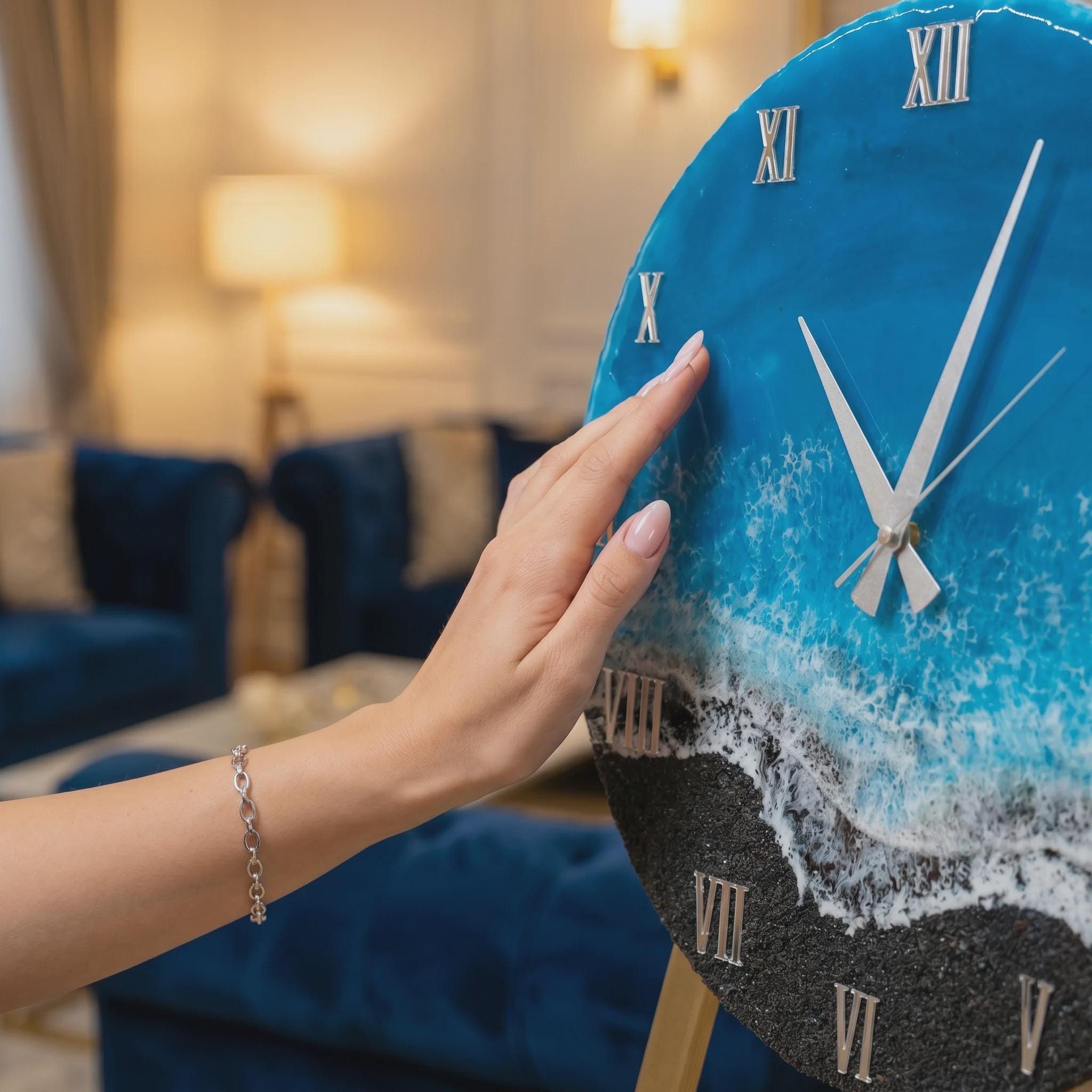 A circular wall clock featuring a resin art design that mimics a beach scene with blue ocean water, white sea foam, and a black sand shoreline. The clock face includes silver Roman numerals and thin silver clock hands.