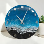 A round wall clock featuring a glossy resin finish that mimics an ocean scene with deep blue water, white sea foam, and a textured black sand beach. The clock face displays silver Roman numerals and two silver clock hands.