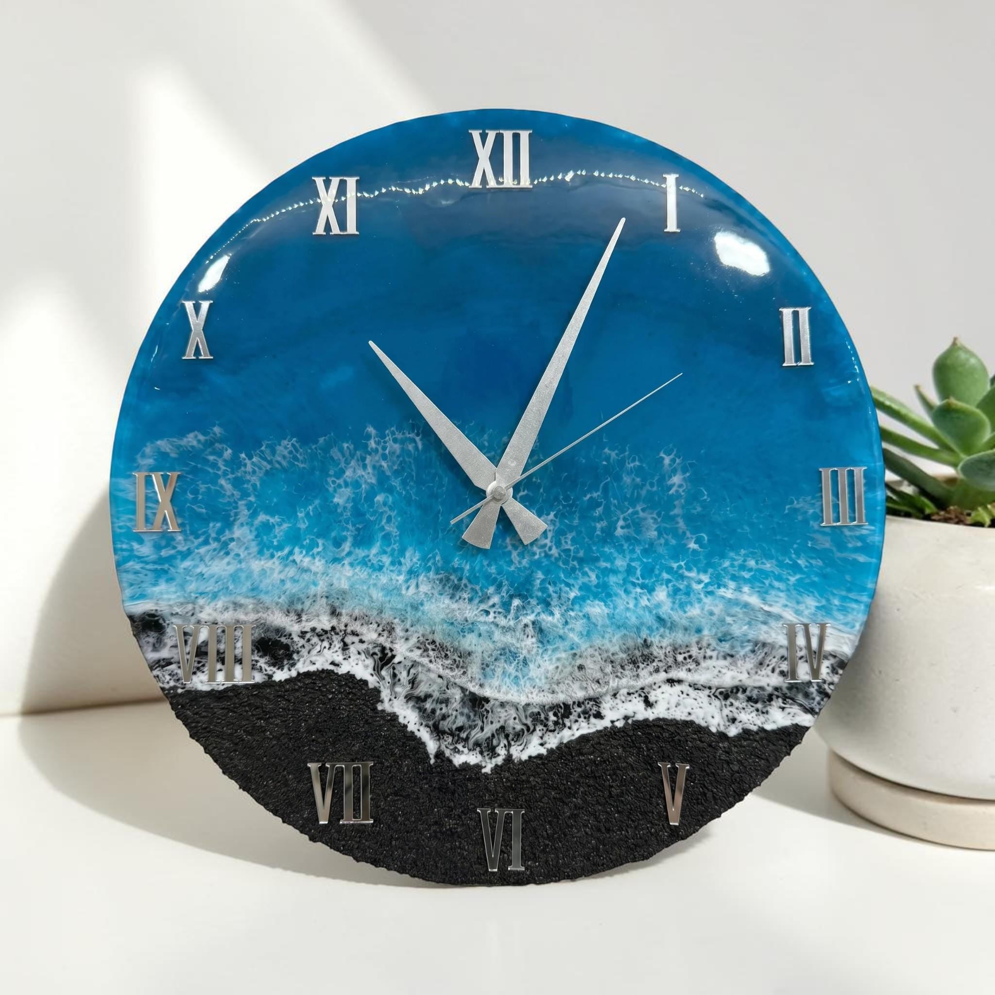A round wall clock featuring a glossy resin finish that mimics an ocean scene with deep blue water, white sea foam, and a textured black sand beach. The clock face displays silver Roman numerals and two silver clock hands.